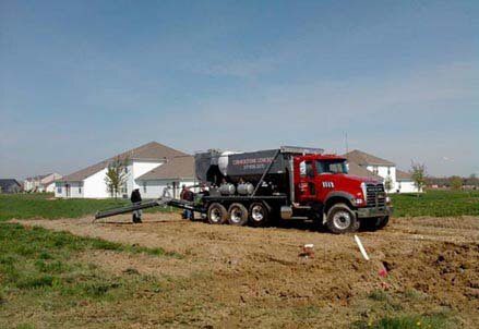 Volumetric concrete mixing truck is on job site, loaded and ready to mix concrete. Concrete company is standing at the chute, preparing for a fresh pour.