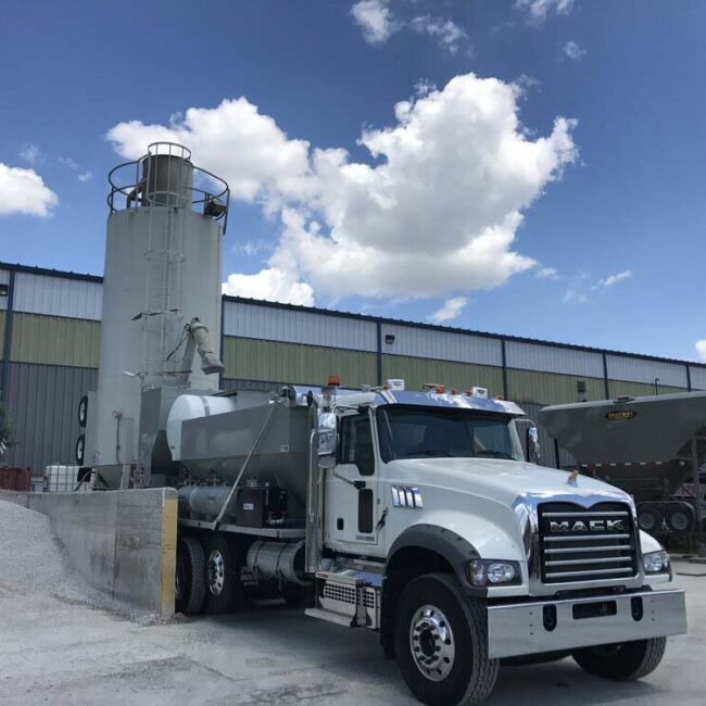 Concrete batching plant. A volumetric concrete mixer is being loaded from a round portable cement silo on job site. A low profile portable silo can be seen in the background.