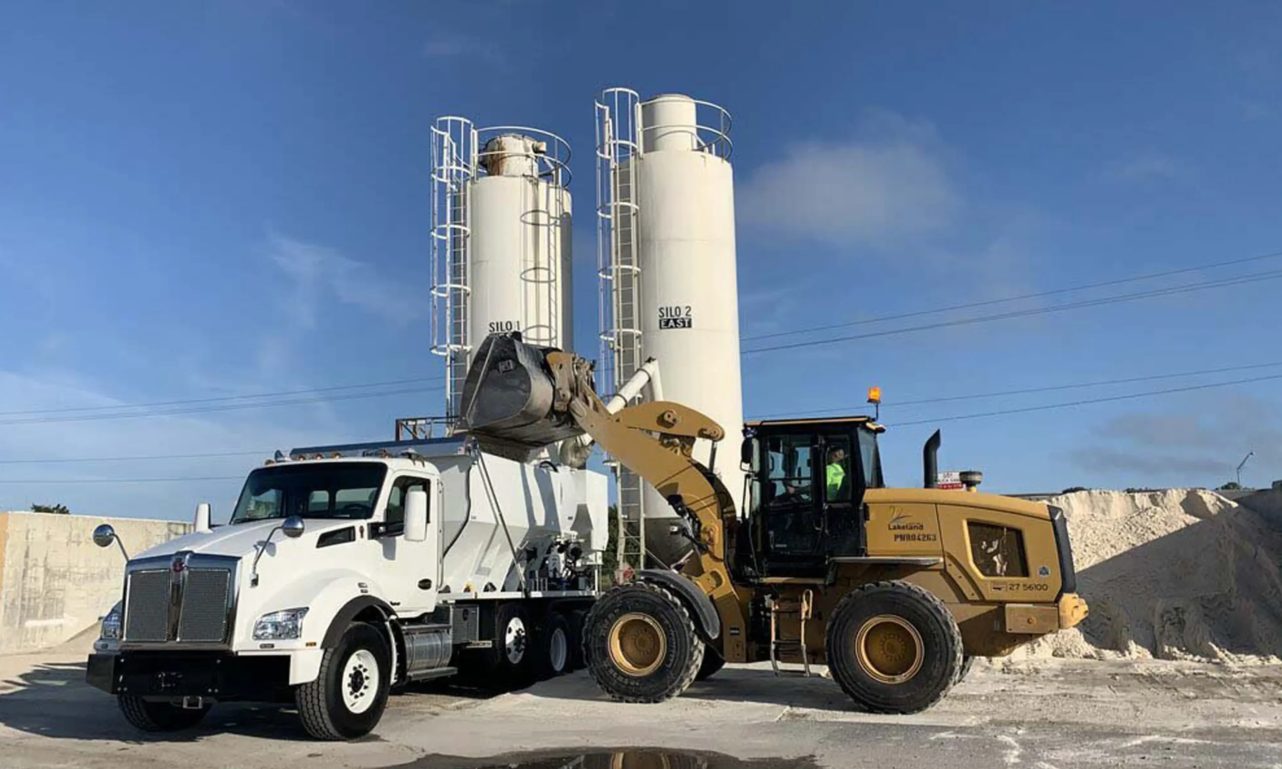 Concrete batching plant - 2 silos behind a volumetric concrete mixer truck being loaded by a front loader. Volumetric concrete mixers can be loaded on the job site for concrete on demand. Cement silos store aggregates and other materials for bulk store and on-site loading.